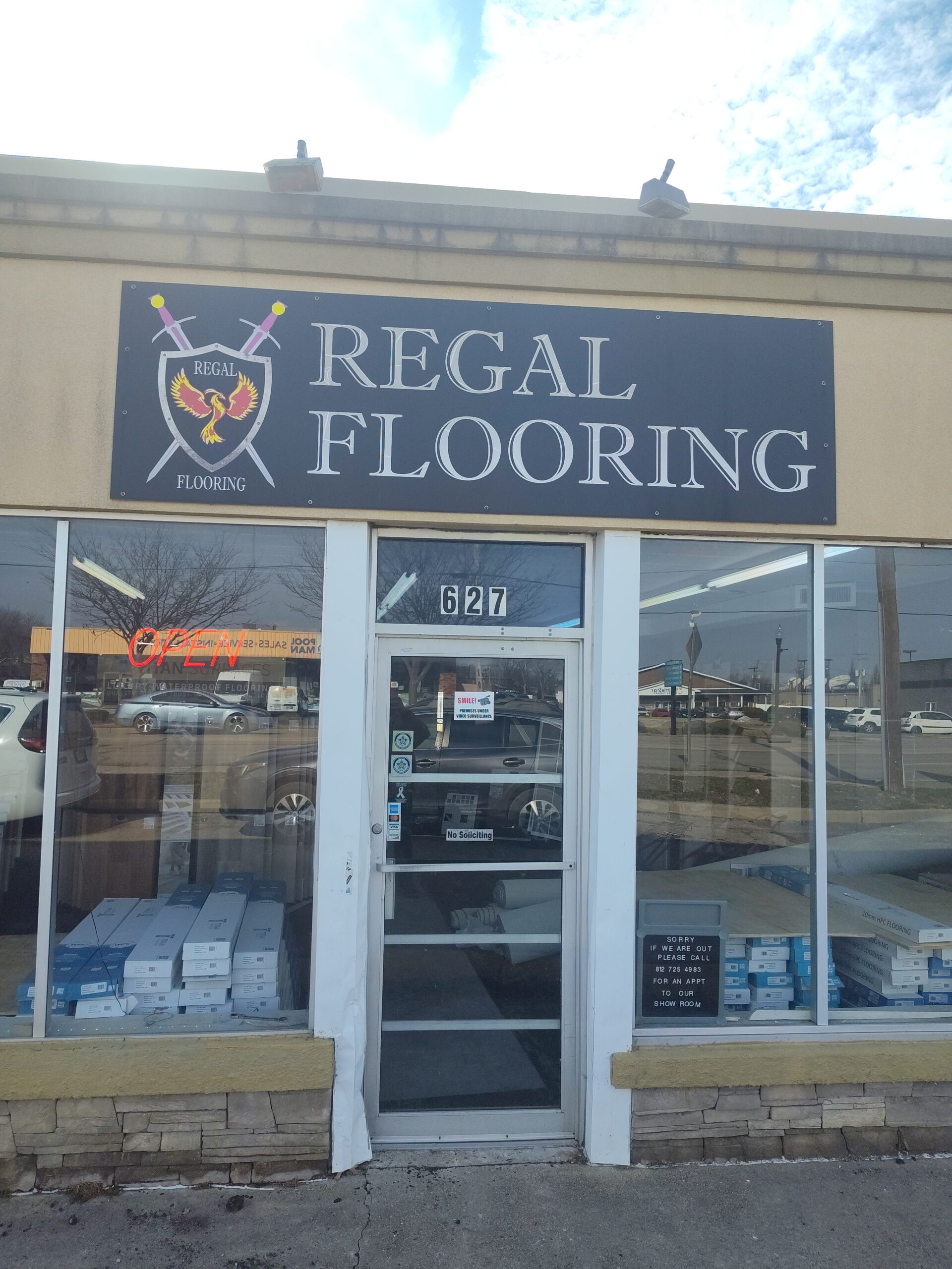 Storefront of Regal Flooring, with a black sign displaying crossed swords and a crest above the entrance. Glass door marked 627, boxes visible inside, and an OPEN sign in the window