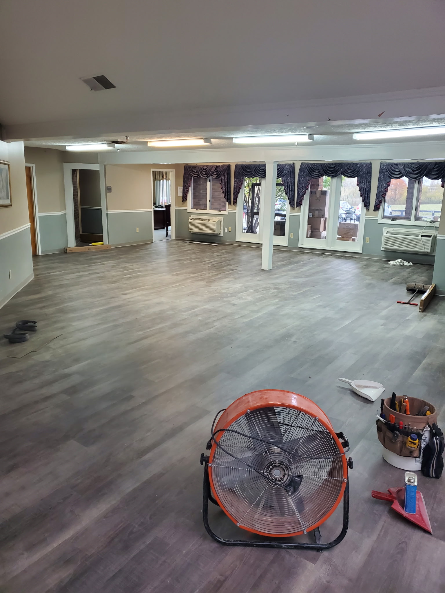 A large, mostly empty room with gray vinyl flooring, a large orange floor fan, and cleaning supplies. Windows with dark curtains line one wall.